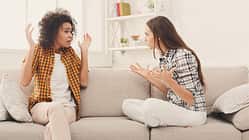 Zwei Frauen sitzen auf einem weißen Sofa und streiten sich. Die linke Frau ist dunkelhäutig, hat braune Locken und trägt ein orangenes Hemd. Die rechte Frau ist weiß, hat braune glatte Haare und trägt ein gemustertes Hemd.