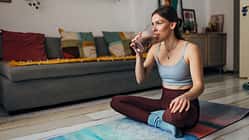 Millennial woman drinks a milkshake while sitting on an exercise mat in the living room