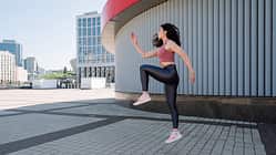 Side view of young woman jumping up, doing cardio workout against urban background.