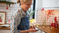 Frau mit Schürze steht in einer weihnachtlich geschmückten Küche und schaut auf ihr Smartphone