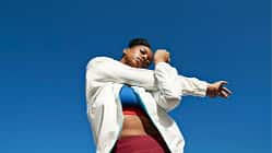 Froschperspektive einer Frau, die ihre Schulter dehnt. Hintergrund nur blauer Himmel