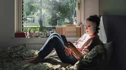 Frau sitzt auf dem Bett am Fenster, trägt eine LED-Maske und schaut auf ihr Smartphone