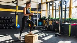 Frau macht Step-Up Übung in Fitnessstudio, im Hintergrund mehrere Fitnessgeräte