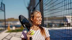 Frau mit Padelschläger und Ball steht lächelnd an einem Metallzaun, Sonnenlicht wirft Gittermuster auf ihr Gesicht.