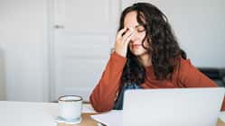 Frau sitzt erschöpft vor einem Laptop, hält sich müde die Stirn, neben ihr stehen eine Tasse und Notizen.