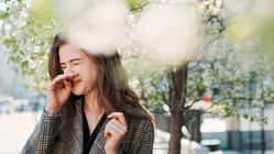 Junge Frau mit Pollenallergie niest draußen unter blühenden Bäumen.