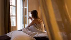 Frau sitzt im Bett mit einem Glas Wasser in der Hand und schaut aus der offenen Balkontür.