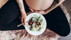 Eine schwangere Frau im Schneidersitz streichelt ihren Bauch und hat einen Teller mit Salat auf dem Schoß