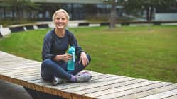 Eine Frau sitzt in Sportkleidung auf einer Parkbank und hält eine Wasserflasche in ihrer Hand