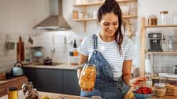 Eine junge Frau in Latzhose steht lächelnd in ihrer Küche und bereitet sich ein Müsli vor