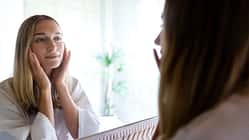 Young blond woman looking at the mirror touching her face. Female examining her facial skin looking in bathroom mirror. Wellness and lifestyle