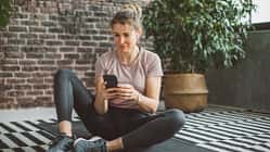 Eine Frau in Trainingskleidung sitzt auf einer Yogamatte und schaut auf ihr Handy