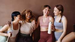 Vier Frauen in Fitnesskleidung stehen lachend vor einem braunen Hintergrund