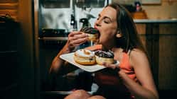 Frau sitzt in der Küche auf dem Boden mit einem Karton mit Donuts in der Hand und isst gierig einen Donut