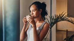 Eine Frau in einem grauen Top schaut aus dem Fenster und hält einen Kaffeebecher in den Händen