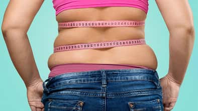 Frau mit sichtbarem Bauchspeck über der Hose wird von hinten mit einem Maßband über dem bauch gezeigt.