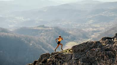 Eine Frau in Laufkleidung läuft einen Bergpass bergauf. Im Hintergrund erstreckt sich ein beeindruckendes Bergpanorama.