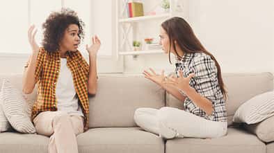 Zwei Frauen sitzen auf einem weißen Sofa und streiten sich. Die linke Frau ist dunkelhäutig, hat braune Locken und trägt ein orangenes Hemd. Die rechte Frau ist weiß, hat braune glatte Haare und trägt ein gemustertes Hemd.