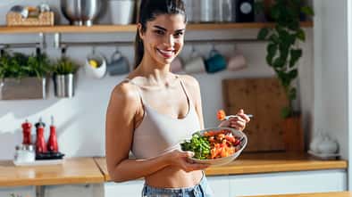 trainierte Frau, die eine gesunde Poke Bowl isst, während sie zu Hause in der Küche in die Kamera schaut