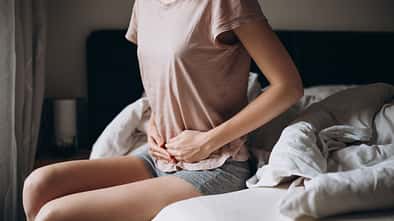 Young woman suffering from strong abdominal pain, casual style indoor shoot. Female sitting on bed in morning Young woman suffering from strong abdominal pain, casual style indoor shoot. Female sitting on bed in morning
