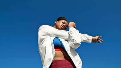 Froschperspektive einer Frau, die ihre Schulter dehnt. Hintergrund nur blauer Himmel