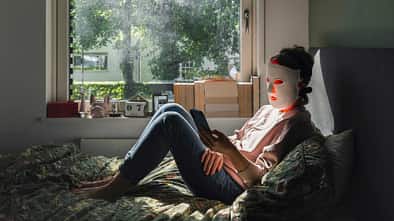 Frau sitzt auf dem Bett am Fenster, trägt eine LED-Maske und schaut auf ihr Smartphone
