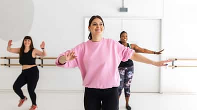 Drei Frauen machen gemeinsam ein Tanz-Workout in einem hellen Fitnessstudio, die Frau vorne trägt ein rosa Shirt und lächelt. Drei Frauen machen gemeinsam ein Tanz-Workout in einem hellen Fitnessstudio, die Frau vorne trägt ein rosa Shirt und lächelt.