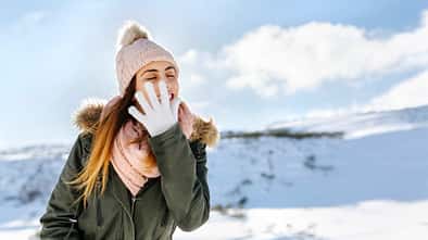 Junge Frau steht lächelnd in einer bergigen Schneelandschaft und hält Hand mit weißem Handschuh vor ihr Gesicht