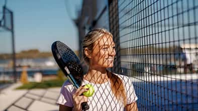 Frau mit Padelschläger und Ball steht lächelnd an einem Metallzaun, Sonnenlicht wirft Gittermuster auf ihr Gesicht.