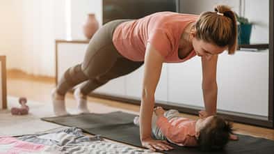 Frau macht eine Plank-Übung auf der Yogamatte, während ihr Baby unter ihr liegt und sie anschaut.