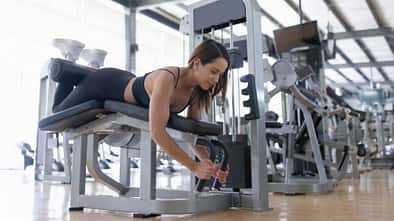 Frau trainiert im Fitnessstudio an einer Beinbeuger-Maschine in Bauchlage und führt eine kontrollierte Kraftübung aus.