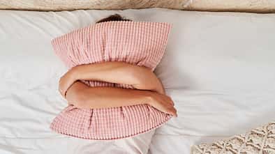 Eine Frau liegt im Bett und verbirgt ihr Gesicht hinter einem Kissen, das sie mit beiden Armen umschlungen hält Eine Frau liegt im Bett und verbirgt ihr Gesicht hinter einem Kissen, das sie mit beiden Armen umschlungen hält