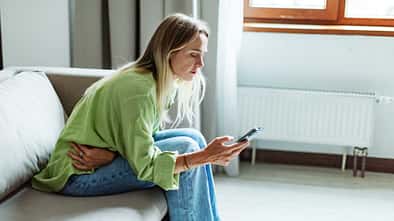 Frau mit Menstruationsbeschwerden sitzt auf dem Sofa und checkt ihre Tracking-App auf dem Smartphone