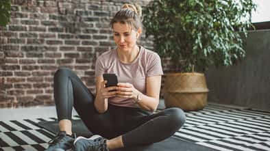 Eine Frau in Trainingskleidung sitzt auf einer Yogamatte und schaut auf ihr Handy