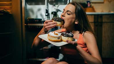 Frau sitzt in der Küche auf dem Boden mit einem Karton mit Donuts in der Hand und isst gierig einen Donut Frau sitzt in der Küche auf dem Boden mit einem Karton mit Donuts in der Hand und isst gierig einen Donut