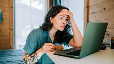 Eine Frau sitzt gestresst nach dem Aufstehen mit einem Kaffee vor ihrem Laptop