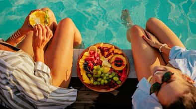 Zwei Frauen sitzen am Pool und essen einen Obstteller