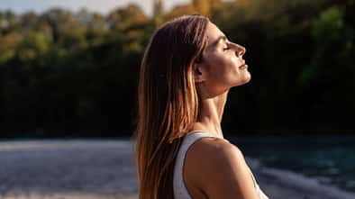 Eine Frau blickt mit geschlossenen Augen in Richtung Himmel. Die Sonne scheint auf ihr Gesicht