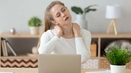Nackenschmerzen, Homeoffice, Rückenschmerzen Frau sitzt am Laptop im Wohnzimmer und reibt sich den schmerzenden Nacken