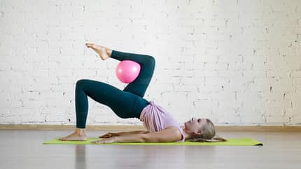 Hip Thrust, Hüftheben Frau hebt aus der Rückenlage die Hüfte an und streckt ein Bein in die Luft. In der Kniekehle des schwebenden Beins hält sie einen Pilates-Ball fest.