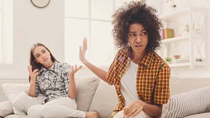 Zwei Frauen sitzen auf einem weißen Sofa und streiten sich. Die linke Frau ist weiß, hat braune glatte Haare und trägt ein gemustertes Hemd. Die rechte Frau ist dunkelhäutig, hat braune Locken und ein orangenes Hemd an. Sie ist abgewandt von der ersten Frau. Zwei Frauen sitzen auf einem weißen Sofa und streiten sich. Die linke Frau ist weiß, hat braune glatte Haare und trägt ein gemustertes Hemd. Die rechte Frau ist dunkelhäutig, hat braune Locken und ein orangenes Hemd an. Sie ist abgewandt von der ersten Frau.