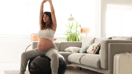Schwangerschaft, Rückenschmerzen Frau mit Babybauch sitzt auf Gymnastikball und streckt sich.