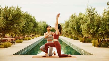 Eine Yoga-Lehrerin zeigt einer jungen Frau eine Yoga-Übung vor einem Pool Eine Yoga-Lehrerin zeigt einer jungen Frau eine Yoga-Übung vor einem Pool