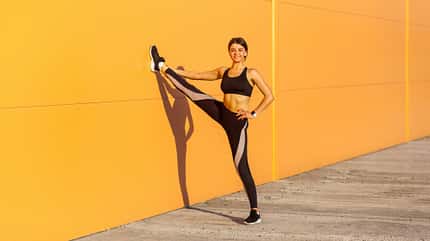 Sportliche Frau trägt schwarze Sportkleidung und übt Sportübungen am Morgen auf der Straße aus auf orangefarbenem Wandhintergrund Sportliche Frau trägt schwarze Sportkleidung und übt Sportübungen am Morgen auf der Straße aus auf orangefarbenem Wandhintergrund