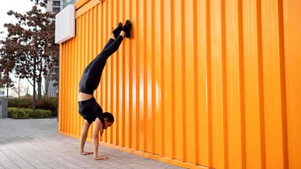 Junge Frau, die im Freien Handstand gegen orangefarbene Wand macht Junge Frau, die im Freien Handstand gegen orangefarbene Wand macht