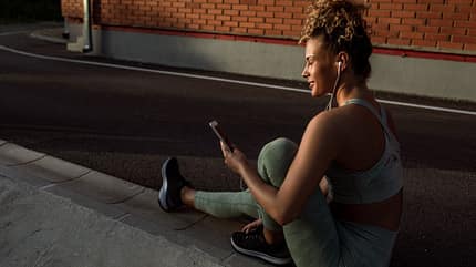 Junge Frau, die ihr Telefon und ihre Ohrhörer nach dem Training im Freien benutzt Junge Frau, die ihr Telefon und ihre Ohrhörer nach dem Training im Freien benutzt