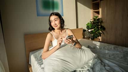 asian and indian ethnicities Woman sitting and stretching on bed, looking out to the city skyline, tapping her smart watch to check notifications