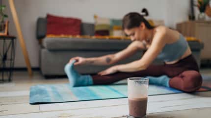 Fit woman is doing stretching exercises at home. Focus on the cup of healthy milkshake that the woman will drink after stretching Fit woman is doing stretching exercises at home. Focus on the cup of healthy milkshake that the woman will drink after stretching