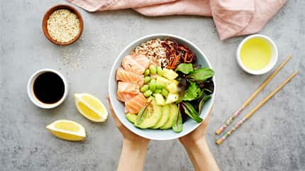 Weibliche Hände halten eine Bowl mit Lachs, Salat, Avocado, edamame Bohnen, Ananas, Sesam und braunem Reis Weibliche Hände halten eine Bowl mit Lachs, Salat, Avocado, edamame Bohnen, Ananas, Sesam und braunem Reis