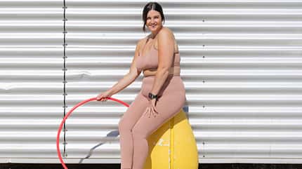 Eine Frau sitzt in Sportkleidung auf einem Hocker und hält einen Hula-Hoop-Reifen